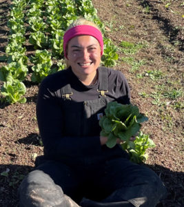 woman seated in field of lettuce, holding a head of lettuce