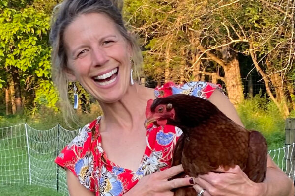a close up image of a smiling white woman with light brown hair holding a brown chicken