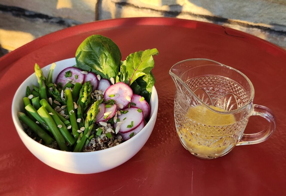 A bowl of fresh salad with asparagus, sliced radishes, leafy greens, and grains topped with seeds, served beside a small glass pitcher of dressing on a red table in warm sunlight.